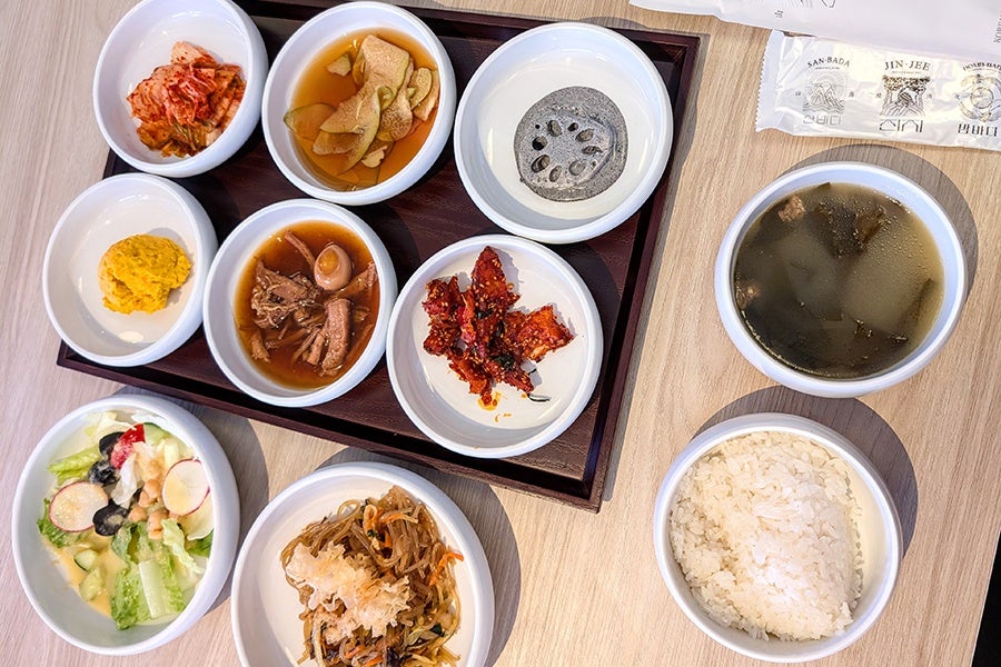 A Korean meal set featuring multiple small white bowls on a wooden tray and table. The tray holds kimchi, pickled radish, lotus root slices, a yellow mashed dish, braised meat with an egg, and spicy dried fish. On the table outside the tray are a bowl of seaweed soup, a bowl of steamed white rice, a mixed vegetable salad with dressing, and a bowl of japchae (stir-fried glass noodles with vegetables).
