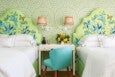 Two beds with green floral and butterfly-patterned headboards flank a white desk with a turquoise upholstered chair. The wall behind features a green geometric pattern, and two wicker wall lamps are mounted above the desk, which holds a vase of yellow flowers, a phone, and a small clock.