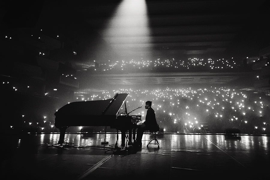 A black and white photo of a musician playing a grand piano on stage, illuminated by a single spotlight. The audience in the background holds up numerous small lights, creating a starry effect in the dark concert hall.