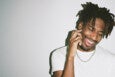 A smiling man with short dreadlocks wearing a white t-shirt and a silver chain necklace, touching his face with one hand against a plain light gray background.
