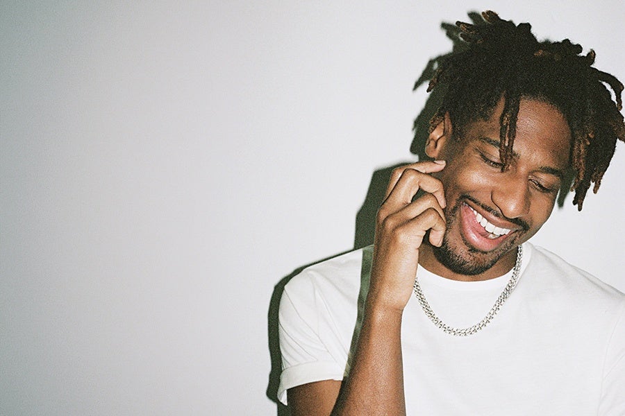 A smiling man with short dreadlocks wearing a white t-shirt and a silver chain necklace, touching his face with one hand against a plain light gray background.