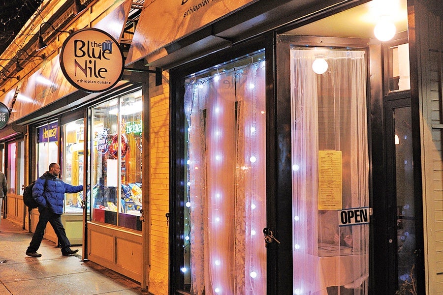 A person wearing a blue jacket and carrying a backpack is opening the door of "The Blue Nile," an Ethiopian cuisine restaurant. The entrance is decorated with white string lights and sheer curtains, and there is an "OPEN" sign on the glass door. The restaurant sign is round and prominently displayed above the door.