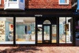 A Sisley Paris storefront with large glass windows displaying a bright, modern interior. Inside, there are makeup stations with chairs, mirrors, and shelves filled with beauty products. The exterior features a black awning with the Sisley Paris logo in white, set against a red brick building. The sidewalk in front is made of red bricks.