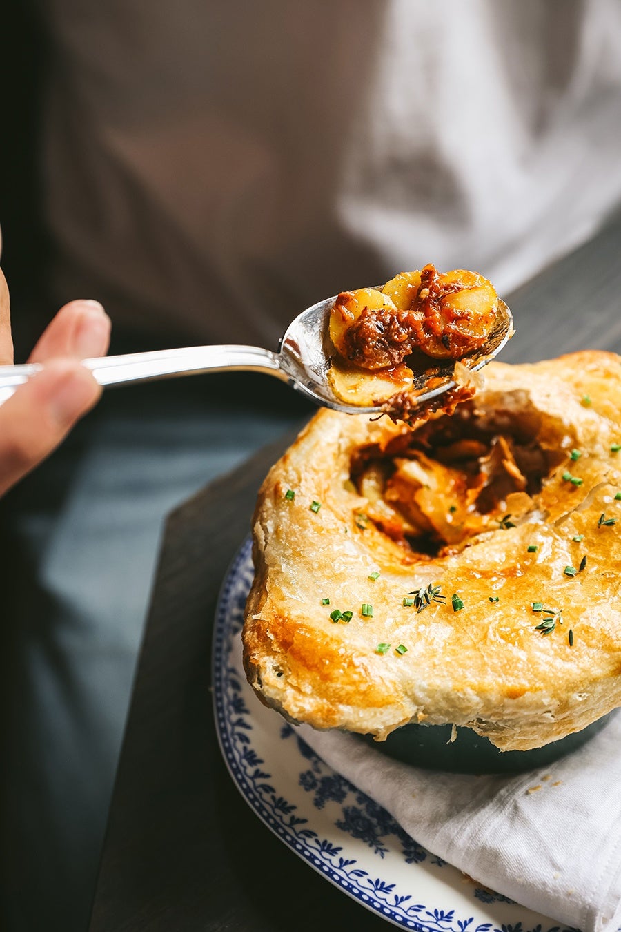Golden-brown pot pie with a flaky crust garnished with chopped herbs, served in a blue and white patterned bowl on a white cloth. A spoon lifting a portion of the filling, which includes sliced potatoes and a rich, savory sauce.