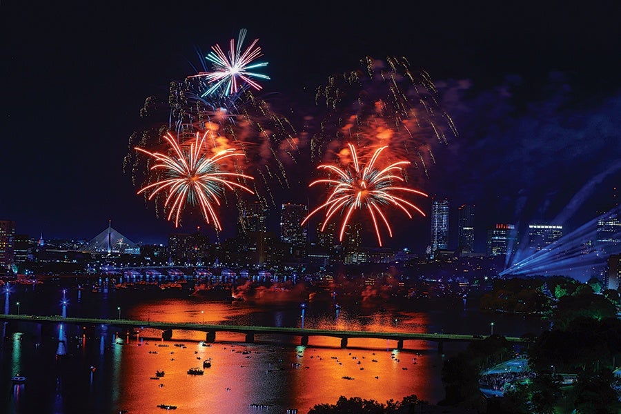 Fireworks bursting in red, white, and blue colors over a city skyline at night, reflecting on a river below with a bridge and boats visible on the water.