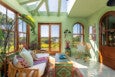 Sunlit living room with light green walls, large windows, and a glass ceiling. The room features a white cushioned wicker sofa with patterned pillows, a colorful floral-patterned coffee table, a wicker rocking chair with a striped pillow, and a small side table with a pink vase of flowers. Wooden framed glass doors and windows offer a view of a lush green landscape and a river. The floor has a light patterned tile with a floral rug underneath the coffee table. Several framed artworks hang on the wall near the rocking chair.