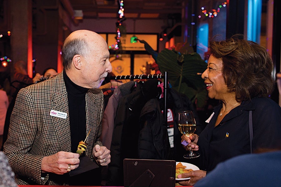 A man and a woman are engaged in conversation at an indoor event. The man, wearing a checkered blazer and black turtleneck, holds a skewer with food and has a name tag on his jacket. The woman, dressed in a dark outfit, holds a glass of white wine and a plate with food. The background features coats hanging on a rack and colorful string lights.