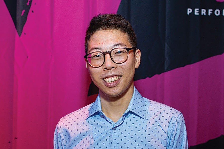 Young man with short dark hair and glasses, smiling, wearing a light blue patterned button-up shirt, standing in front of a pink and black background.