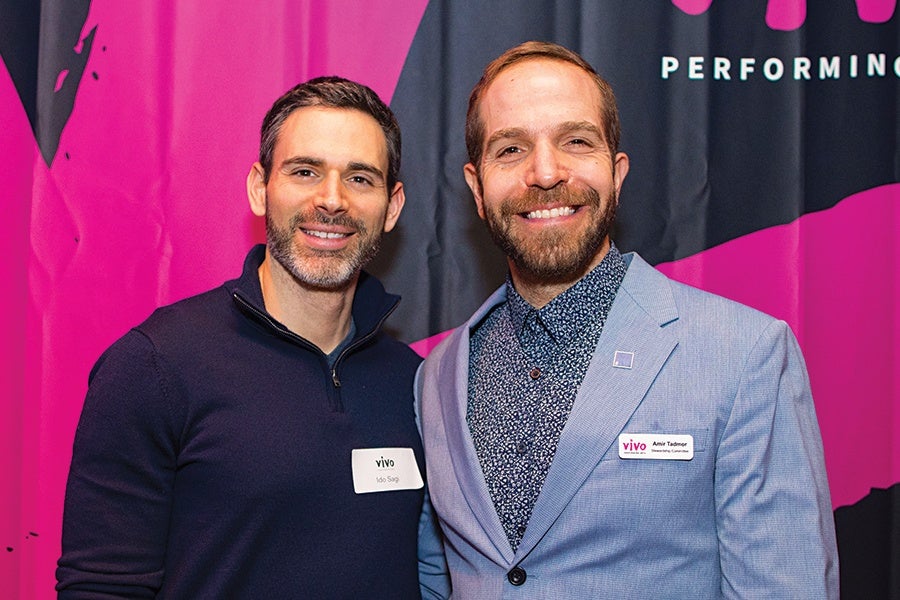 Two men smiling and posing together in front of a pink and black backdrop. The man on the left is wearing a dark navy quarter-zip sweater with a name tag that reads "Ido Sagy." The man on the right is wearing a light blue blazer over a patterned shirt with a name tag that reads "Amir Tadmor.