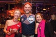 Three people smiling and posing together in a warmly lit indoor setting. The person on the left is wearing a red one-shoulder dress and holding a copper mug with a straw. The person in the middle is wearing a colorful patterned shirt with a red beanie. The person on the right is wearing a black outfit with a bright pink scarf. The background shows blurred lights and other people.