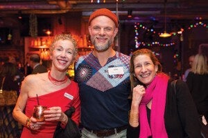 Three people smiling and posing together in a warmly lit indoor setting. The person on the left is wearing a red one-shoulder dress and holding a copper mug with a straw. The person in the middle is wearing a colorful patterned shirt with a red beanie. The person on the right is wearing a black outfit with a bright pink scarf. The background shows blurred lights and other people.
