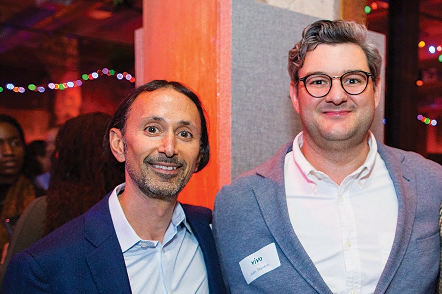Two men are standing side by side indoors with colorful string lights in the background. The man on the left has medium-length dark hair, a trimmed beard, and is wearing a dark blue blazer over a light blue shirt. The man on the right has short, wavy hair, glasses, and is wearing a gray blazer over a white shirt with a name tag that reads "vivo John Stalman." Both are smiling.