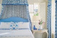 A bedroom featuring a bed with a blue upholstered headboard and white bedding embroidered with blue patterns. The bed has a canopy with blue and white floral fabric, matching the curtains on the window. A small wooden side table next to the bed holds a white vase with pink flowers, two blue glass bottles, a small blue cup, and a book. A white lamp with a beige shade is positioned behind the vase. The room has white walls and windows with white shutters.