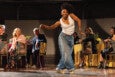 A woman with curly hair wearing a white sleeveless top and loose blue jeans is dancing on a stage. She is smiling and posing with her arms bent and extended to the sides. In the background, several people are seated at tables covered with tablecloths, watching her performance. The setting appears to be an indoor event or gathering.