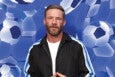 A man with short brown hair and a beard is wearing a black jacket with blue and white stripes on the sleeves over a white shirt. The background features a pattern of blue and white soccer balls.
