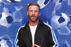 A man with short brown hair and a beard is wearing a black jacket with blue and white stripes on the sleeves over a white shirt. The background features a pattern of blue and white soccer balls.