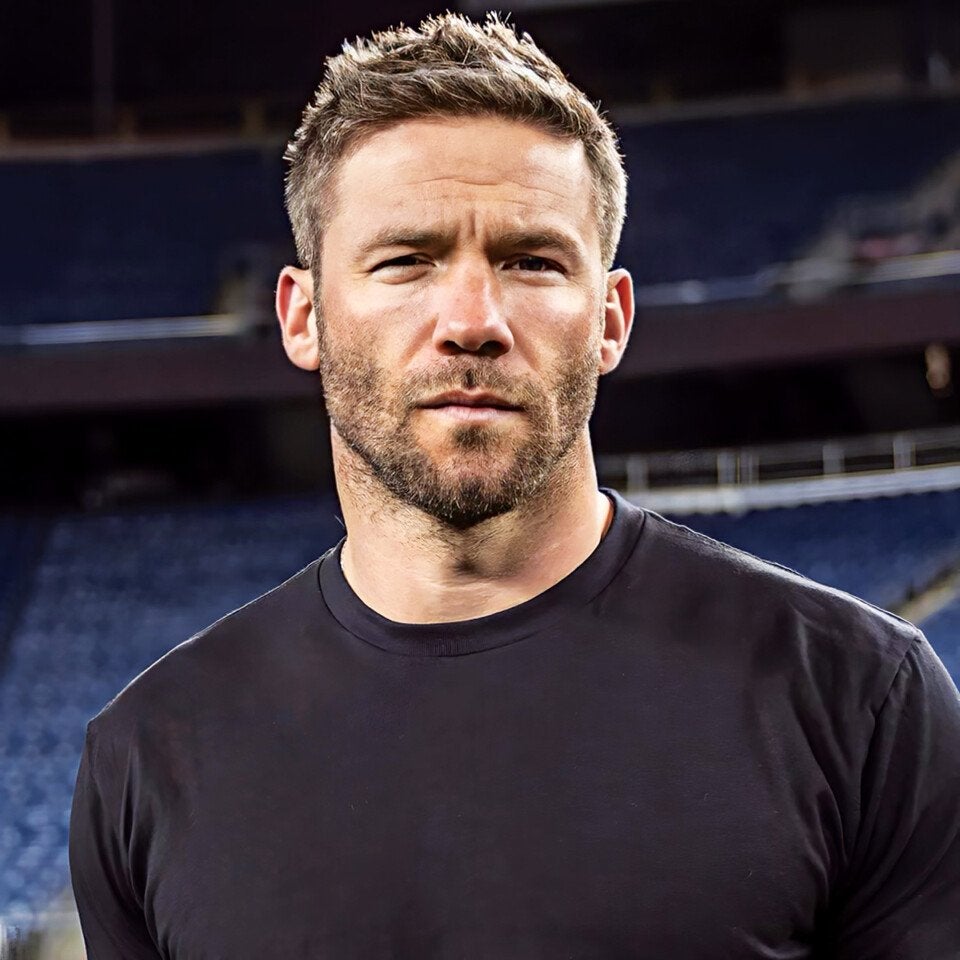 Man with short, light brown hair and a trimmed beard, wearing a plain black t-shirt, standing in front of a blurred background that appears to be a stadium or arena seating area.