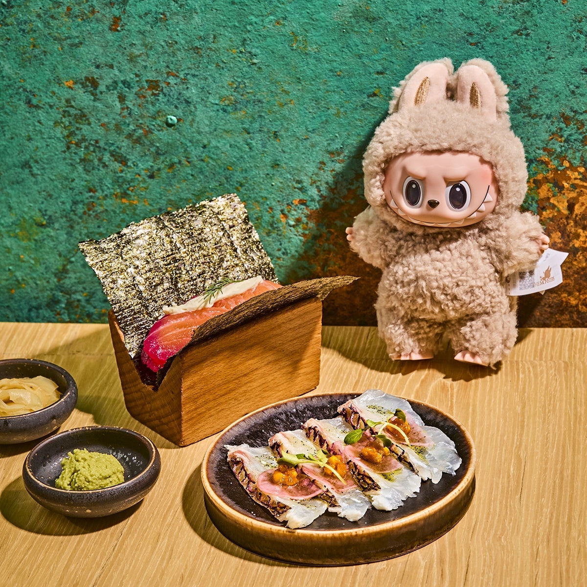 A wooden table holds a black plate with four pieces of sushi topped with garnishes, a wooden box containing seaweed-wrapped sushi with salmon, and two small black bowls, one with green wasabi and the other with pickled ginger. To the right, there is a plush toy with a fluffy beige bunny costume and an expressive face. The background is a textured teal and rust-colored wall.