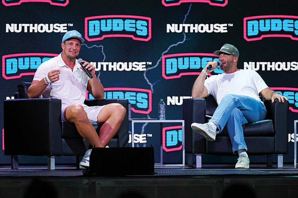Two men are seated on black armchairs on a stage, each holding a microphone. The man on the left wears a light blue cap, white polo shirt, and white shorts, while the man on the right wears a dark green cap, white t-shirt, and blue jeans. A small table with two water bottles is between them. The background features a large screen with the words "NUTHOUSE" and "DUDES" in bold letters.