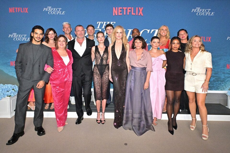 A group of fifteen people posing together at a Netflix event for "The Perfect Couple." They are dressed in a variety of formal and semi-formal outfits, including suits, dresses, and jumpsuits, standing in front of a blue backdrop with the Netflix logo and "The Perfect Couple" title repeated. The setting appears to be an indoor venue with a carpeted floor and floral arrangements on either side.