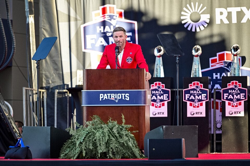 A man in a red blazer with a "Hall of Fame" patch stands behind a wooden podium with a "Patriots" sign, speaking into a microphone. The background features multiple "Hall of Fame" signs and trophies, with the RTX logo visible. Green plants are placed in front of the podium.