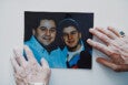 A color photograph of two smiling men, one wearing a light blue shirt and the other wearing a dark jacket and a cap,...