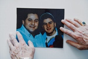 A color photograph of two smiling men, one wearing a light blue shirt and the other wearing a dark jacket and a cap, held on a white surface by two elderly hands. The right hand has a ring with a large dark stone, and the left wrist has a silver watch.