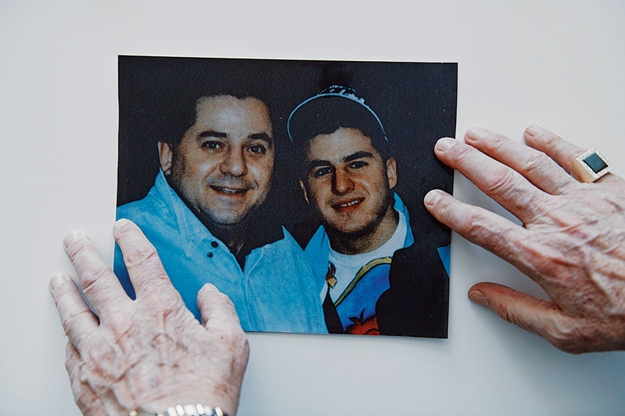 A color photograph of two smiling men, one wearing a light blue shirt and the other wearing a dark jacket and a cap, held on a white surface by two elderly hands. The right hand has a ring with a large dark stone, and the left wrist has a silver watch.