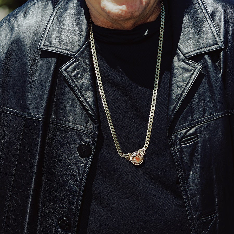 A person wearing a black leather jacket and a black shirt, with a gold chain necklace featuring a round pendant with a reddish-brown stone.