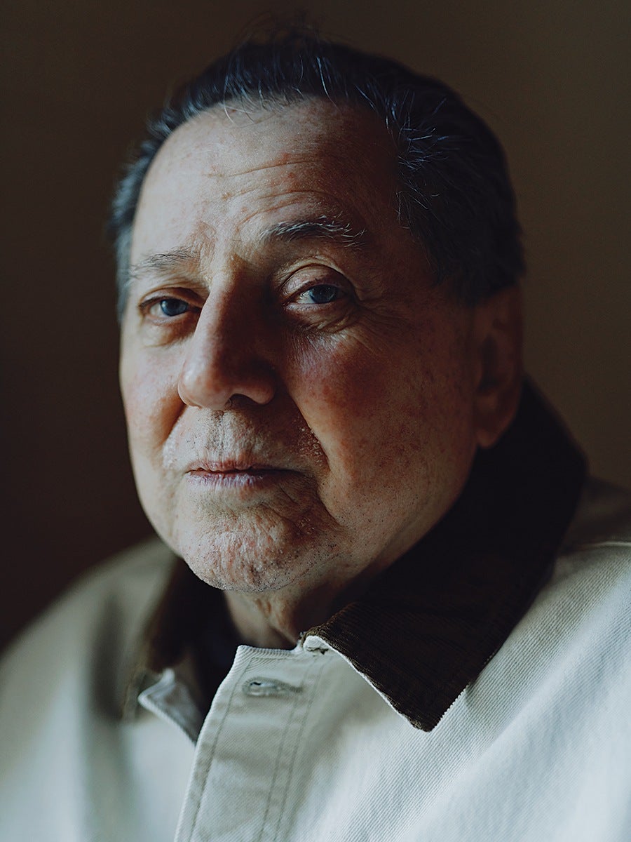 Close-up portrait of an older man with light skin and blue eyes, showing detailed facial features and wrinkles. He has short, dark hair with some gray strands and is wearing a light-colored jacket with a dark collar. The background is dark and out of focus, emphasizing his face.