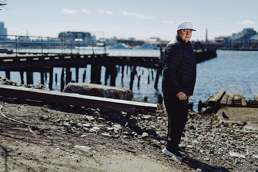A man wearing a white baseball cap, black jacket, black pants, and white sneakers stands on a rocky shoreline near a wooden pier extending over the water. The background includes a body of water with boats and buildings visible in the distance under a clear sky.
