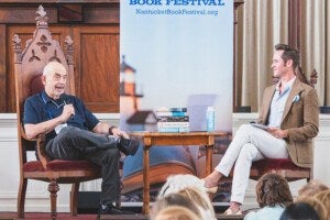 Two men are seated on ornate wooden chairs on a stage, engaged in a discussion at the Nantucket Book Festival. A small wooden table between them holds a stack of books and a water bottle. The man on the left wears a dark polo shirt and dark pants, holding a microphone, while the man on the right wears a light blue shirt, beige blazer, and white pants, holding papers. An audience is partially visible in the foreground.