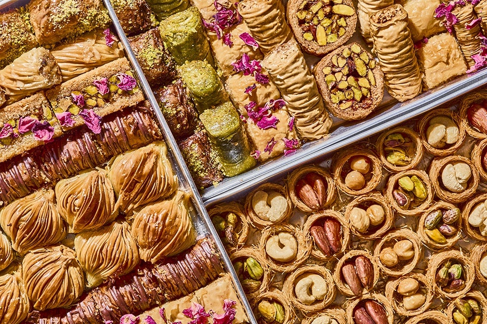 An assortment of Middle Eastern pastries arranged in metal trays. The pastries include various types of baklava and nut-filled sweets, featuring layers of flaky golden-brown phyllo dough, pistachios, cashews, and other nuts. Some pastries are decorated with pink rose petals, adding a colorful and elegant touch. The display showcases a variety of shapes and textures, from rolled and layered to round and nut-topped.