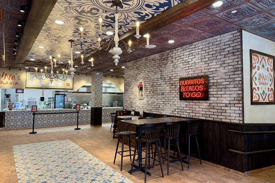 A restaurant interior with a patterned tile floor and ceiling, featuring two ornate chandeliers. The counter area has a tiled front and a menu displayed above. A brick wall on the right side has a neon sign reading "BURRITOS & TACOS TO GO" and a framed colorful sign with the text "Anna's Taqueria." There is a high table with six wooden bar stools in front of the brick wall. The overall decor combines rustic and decorative elements.