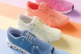Four pairs of athletic shoes in pastel colors are arranged diagonally on a multicolored background. The shoes are in blue, off-white, coral, and pink, each featuring a mesh upper, white soles with a segmented design, and matching laces. The background consists of horizontal stripes in light blue, cream, orange, and lavender.