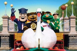 A large white swan paddle boat with a golden handle is in the foreground. Behind it, four sports mascots and a person in a green outfit stand on a dock. From left to right: a mascot in a blue Patriots costume waving, a bear mascot in a black Bruins jersey, a green frog-like mascot in a Red Sox jersey holding a hot dog, and a person in a green vest with shamrocks spinning a basketball on one finger. The background shows a stone bridge with lamp posts and greenery.
