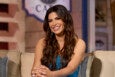 A woman with long, wavy dark brown hair is sitting on a beige couch with blue and beige pillows. She is wearing a sparkly blue strapless dress and silver earrings, smiling with her hands clasped in front of her. The background is softly blurred with warm tones.