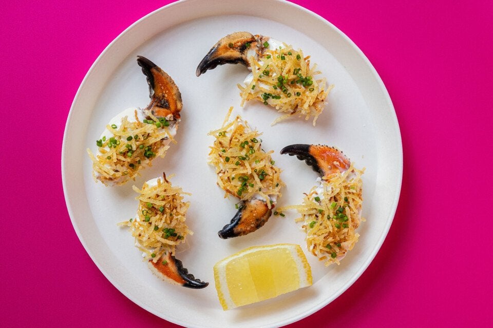 Five crab claws topped with a creamy mixture, crispy shredded potatoes, and chopped chives arranged on a white plate with a lemon wedge, set against a bright pink background.
