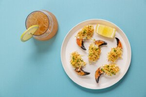 Five crab claws topped with a creamy mixture and crispy shredded potatoes, garnished with chopped chives, served on a white plate with a lemon wedge. Next to the plate is a glass of iced beverage garnished with a lemon slice, all set on a light blue surface.