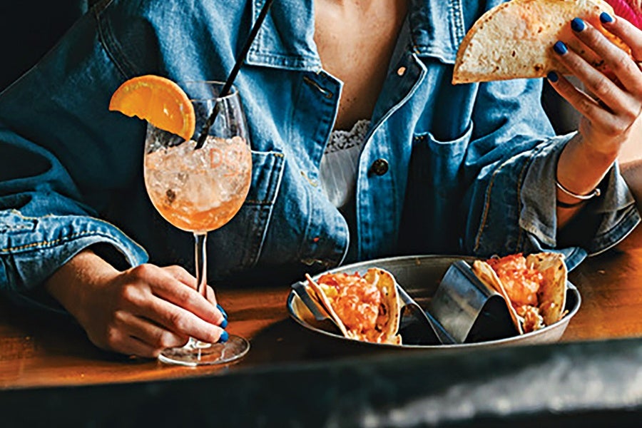 A person wearing a denim jacket is holding a taco in one hand and a wine glass with an orange slice garnish and a black straw in the other. On the table in front of them is a metal tray with two tacos held upright in a taco holder.