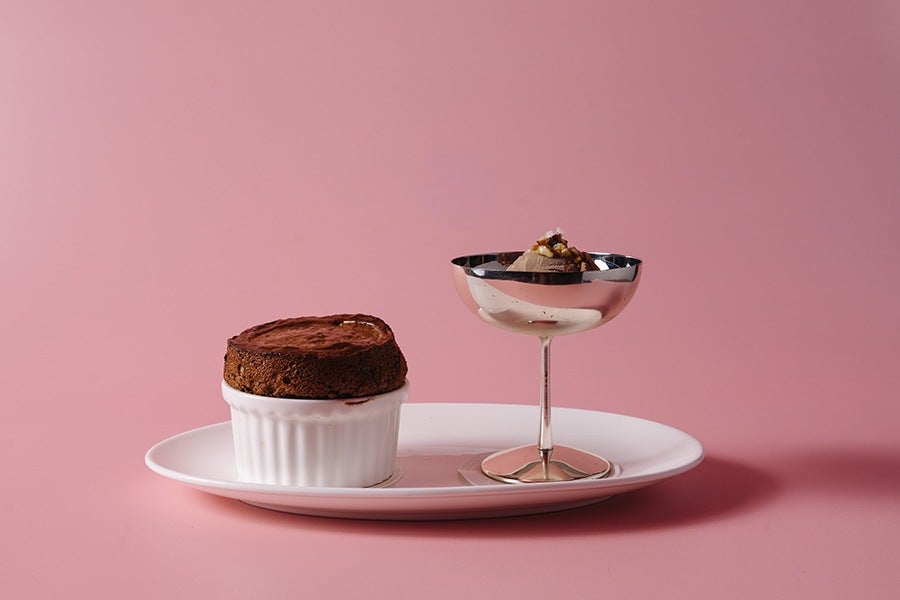 Chocolate soufflé in a white ramekin alongside a silver dessert glass with chocolate mousse, both placed on a white oval plate against a pink background.