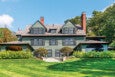 Large green two-story house with multiple windows, two prominent chimneys, and a gray shingled roof. The house is surrounded by well-maintained bushes and trees, with a wide green lawn in the foreground under a clear blue sky.