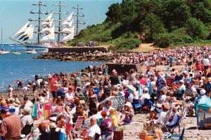 A large crowd of people gathered on a rocky beach, many sitting on folding chairs and some standing or walking. In the background, a tall sailing ship with multiple masts and white sails is sailing near the shore. The scene is set on a sunny day with clear blue skies and green trees on a hill behind the beach. Several smaller boats are also visible on the water.