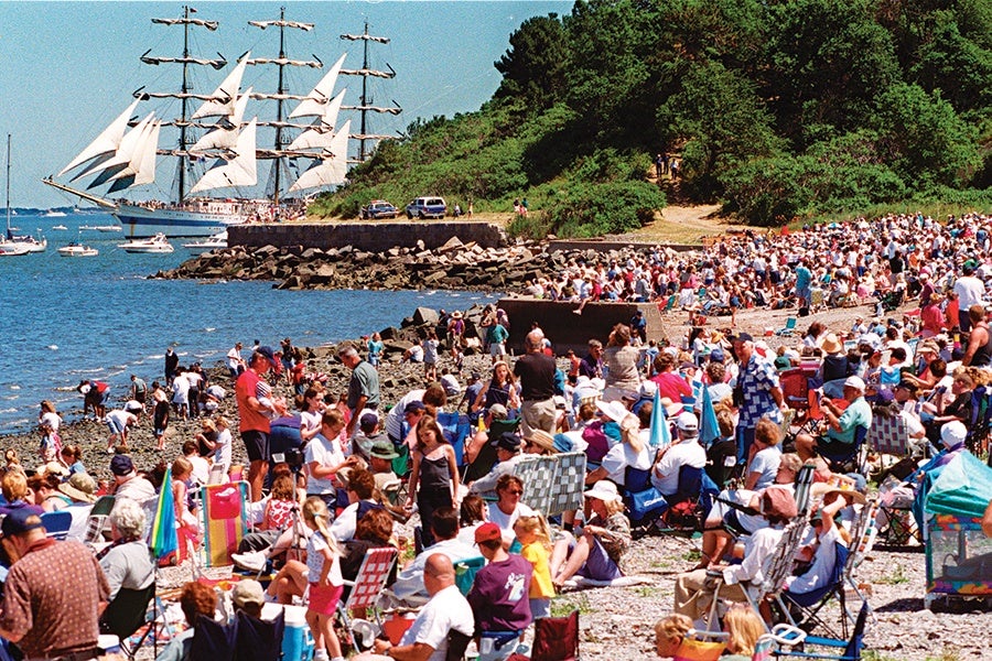 A large crowd of people gathered on a rocky beach, many sitting on folding chairs and some standing or walking. In the background, a tall sailing ship with multiple masts and white sails is sailing near the shore. The scene is set on a sunny day with clear blue skies and green trees on a hill behind the beach. Several smaller boats are also visible on the water.