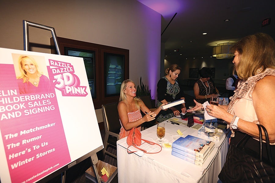 A woman with blonde hair in a sleeveless orange dress is seated at a table signing books. The table is covered with a white cloth and has several stacks of books, a glass of iced tea, and some papers. Another woman with brown hair, wearing a sleeveless light pink top with ruffles, is standing across the table holding a glass and a piece of paper. Behind the seated woman, two other women are organizing items on the table. A large sign on an easel next to the table features a photo of the blonde woman and text promoting a book sales and signing event for "Razzle Dazzle 3D Pink" by an author named Eline Hilderbrand, listing titles such as "The Matchmaker," "The Rumor," "Here's To Us," and "Winter Storms.