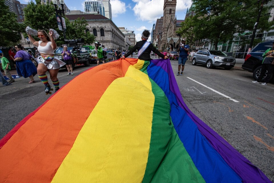 A person wearing a black jacket and a sash holds a large rainbow pride flag that stretches across a city street during a parade. To the left, a woman in a white crop top, silver skirt, and rainbow knee-high socks roller skates while holding silver pom-poms. Several people and vehicles are visible in the background, along with tall buildings and trees lining the street.