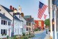 A small town street lined with white and light-colored wooden houses featuring black shutters and American flags hanging from the buildings. A tall streetlamp is lit, and a clock tower with a golden dome is visible in the background. The street curves slightly and leads to a red brick building further down. The sky is clear and blue.