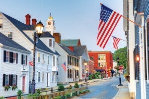 A small town street lined with white and light-colored wooden houses featuring black shutters and American flags hanging from the buildings. A tall streetlamp is lit, and a clock tower with a golden dome is visible in the background. The street curves slightly and leads to a red brick building further down. The sky is clear and blue.