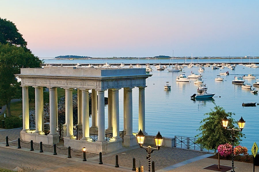 A classical-style stone pavilion with tall columns is situated near a waterfront. The calm water is dotted with numerous small boats, and the sky shows soft pastel colors of early evening or dawn. Street lamps and trees are visible along the paved walkway beside the pavilion.
