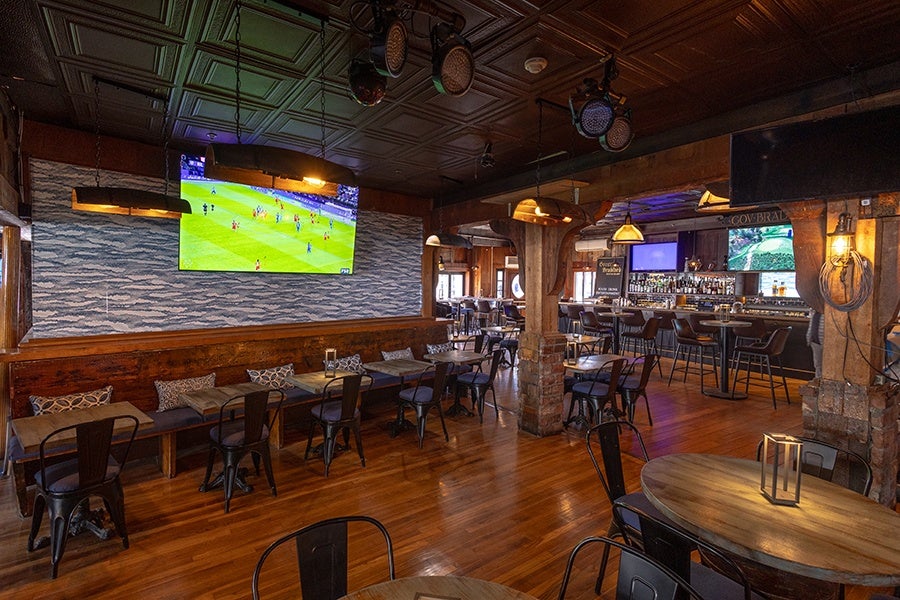 Wood-floored bar and dining area with wooden tables and black metal chairs. A long wooden bench with patterned cushions lines one wall beneath a large flat-screen TV showing a soccer game. The bar area features high stools and multiple TV screens, with warm pendant lighting and exposed brick columns adding rustic charm.